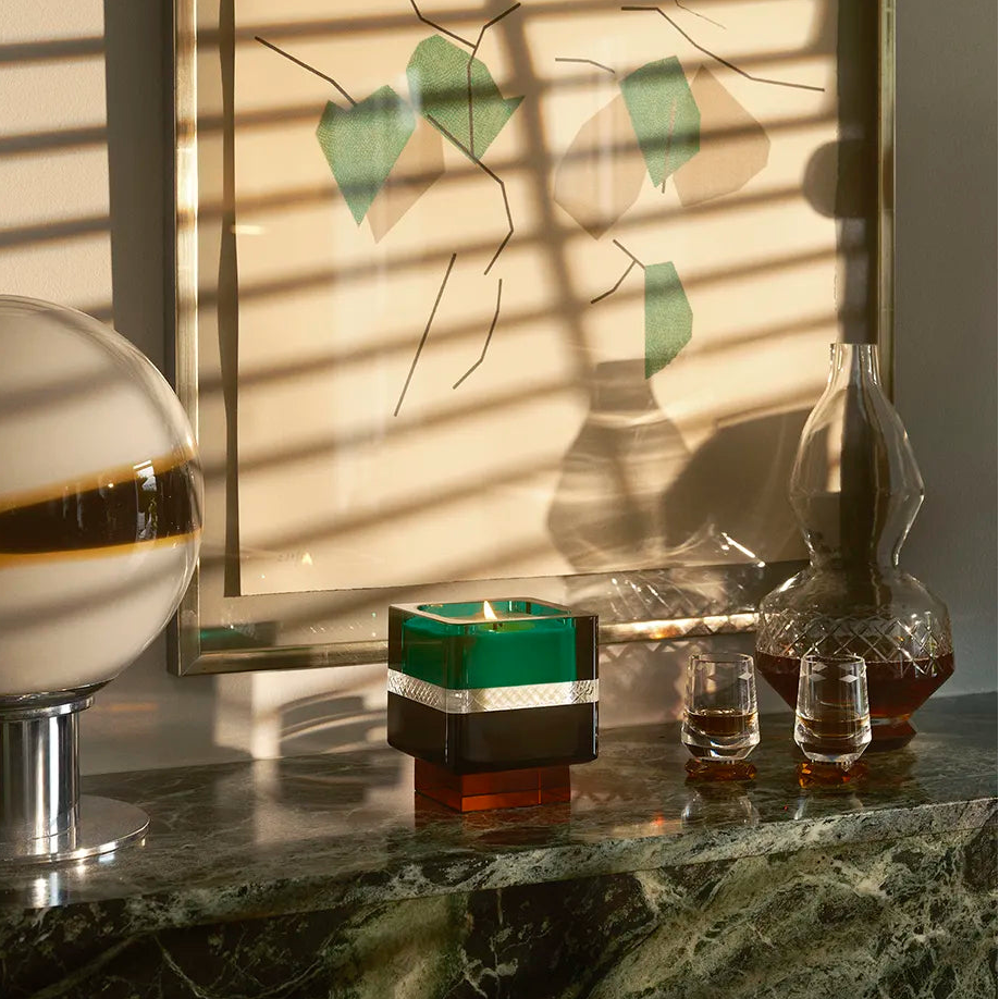 The Soirée Home Scent candle rests on a marble shelf, surrounded by crystal glassware, with sunlight casting striking shadows across the scene.