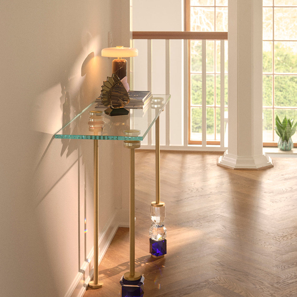 A modern console table with a clear glass top and slim gold-toned metal legs, styled with a lamp and books in a bright interior hallway with wood floors and large windows.