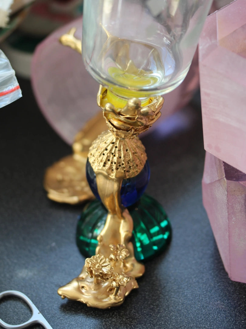 A close-up view of the "Adaptation Glass," showcasing its intricate design. The crystal goblet features an 18-carat gold-plated silver stem, adorned with a shell motif and flowing gold detailing. Vibrant emerald green and cobalt blue accents, along with floral embellishments, add a luxurious touch to the artistic structure.