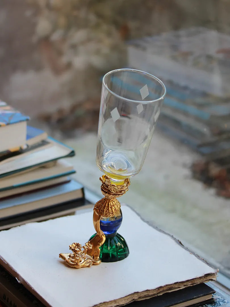 A uniquely crafted goblet titled "Adaptation Glass," made from crystal and 18-carat gold-plated silver. The stem features a vibrant combination of emerald green, cobalt blue, and gold detailing, shaped like an artistic sculpture with a textured gold accent resembling a seashell and floral embellishments near the base. The glass rests on a stack of books with a blurred background.