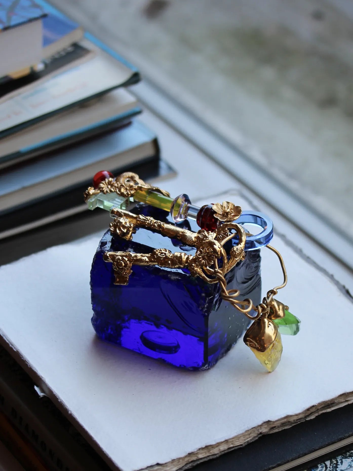 A decorative object titled "Bloom," featuring a vibrant blue crystal cube adorned with 18-carat gold-plated silver details. The design includes intricate floral and vine motifs in gold, along with colorful crystal accents in red, green, and yellow. Hanging charms add movement and additional elegance to this artistic piece, displayed on a stack of books.