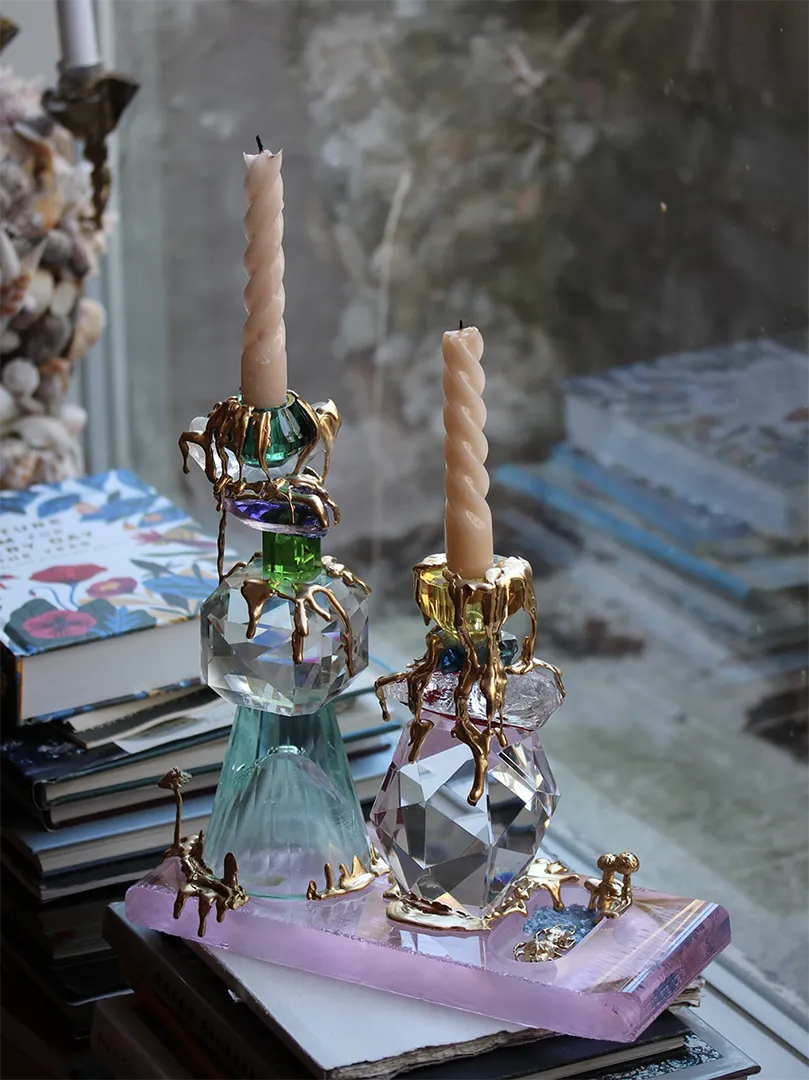 A pair of luxurious candle holders titled "Metamorphosis," crafted from faceted crystal and 18-carat gold-plated silver. Each holder features a dynamic combination of rose, green, clear, blue, and purple crystal elements. The design includes flowing gold accents that mimic molten metal, elegantly draped over the crystals. Topped with spiral beige candles, the holders rest on stacked books near a softly lit window.