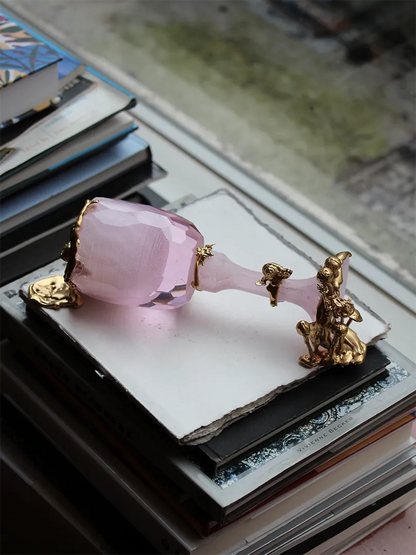 "Symbiosis," a decorative object crafted from rose-colored crystal and 18-carat gold-plated silver, features a striking design. The faceted crystal is paired with gold sculptural elements, including a small figure and natural textures, symbolizing the harmony of art and nature. Displayed on a pile of books with a rustic backdrop.