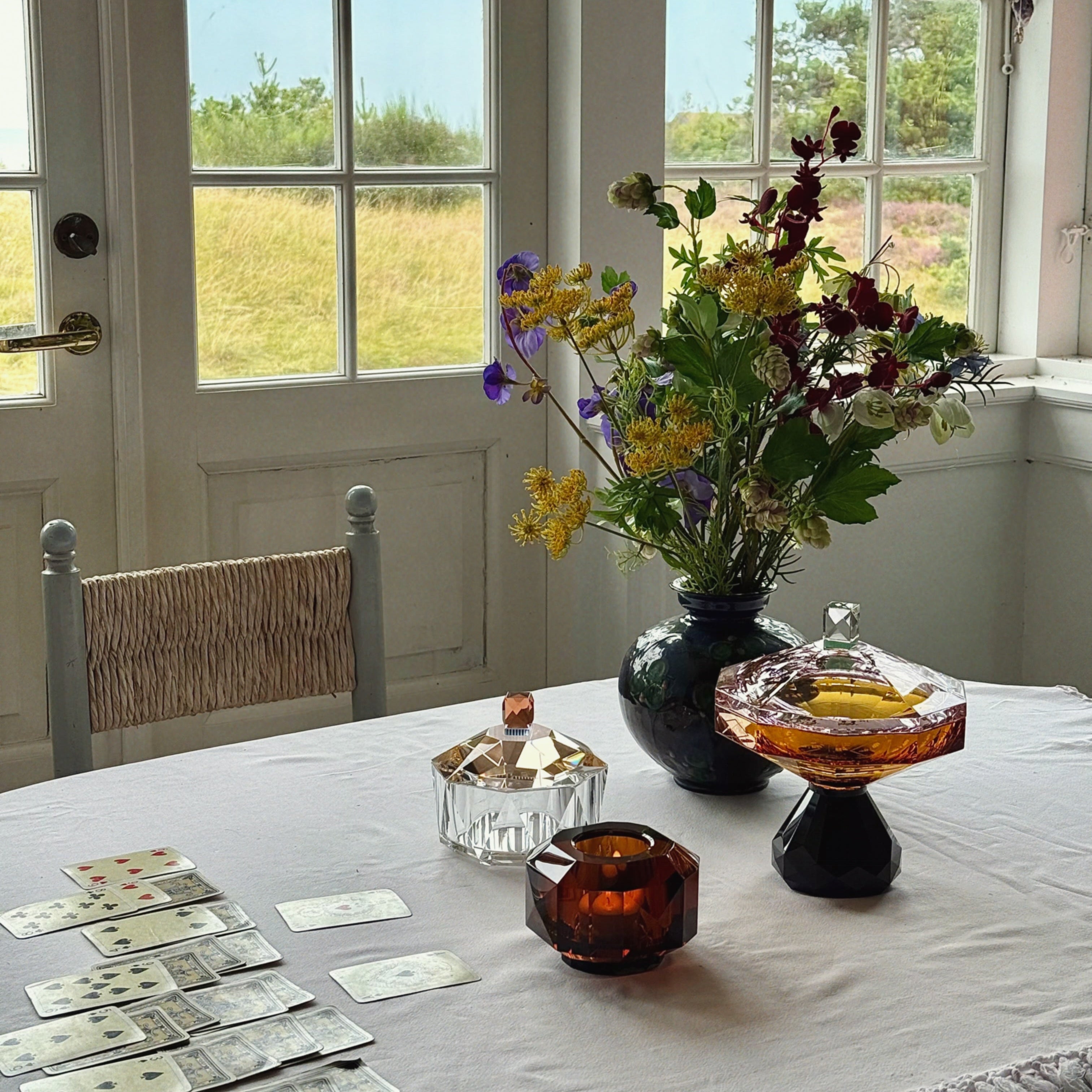 A styled dining table in a bright room with large white-framed windows overlooking a grassy outdoor field. On the table are several playing cards, a dark vase filled with mixed flowers, a faceted amber glass container, and a clear glass container with a gold lid. Reflections Copenhagen