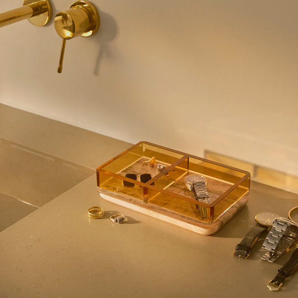 The Treasures Tray with an amber crystal cover and onyx base sits on a bathroom counter, elegantly organizing watches and jewelry in golden light.