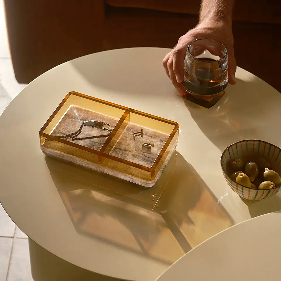 A Treasures Tray with a warm amber glow rests on a coffee table, holding accessories, as a hand reaches for a whisky glass nearby.