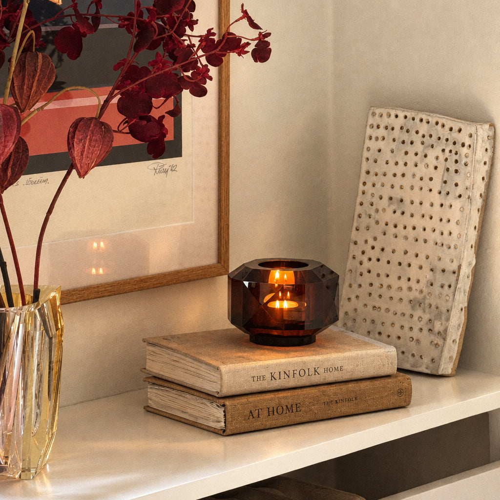A lit amber crystal candle holder with a faceted geometric design sits on a stack of two books on a white shelf. The shelf is styled with a gold vase containing dark red flowers, a framed print, and a white textured decorative board. Copenhagen, Andrea Larsson